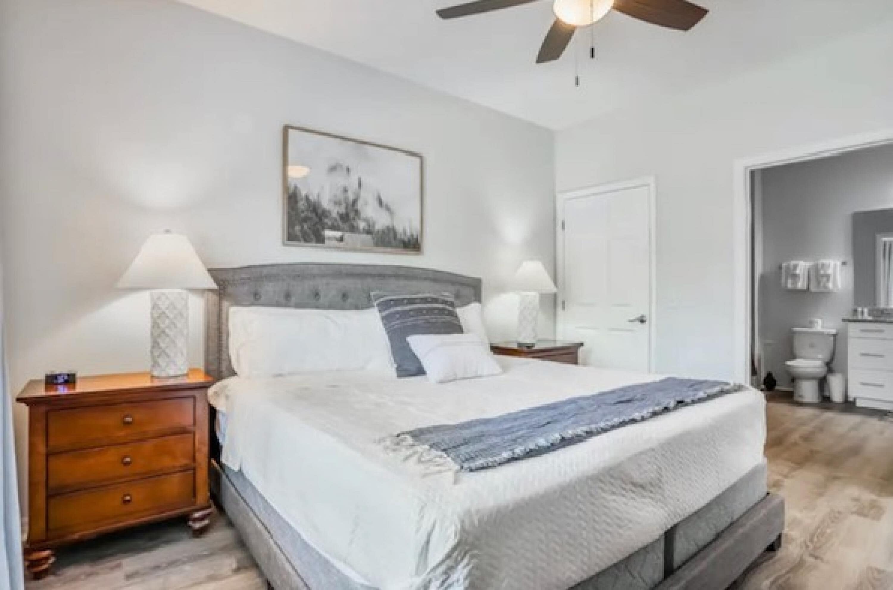 A bedroom with a bed, nightstands, a ceiling fan, a framed picture, and a door leading to a bathroom.