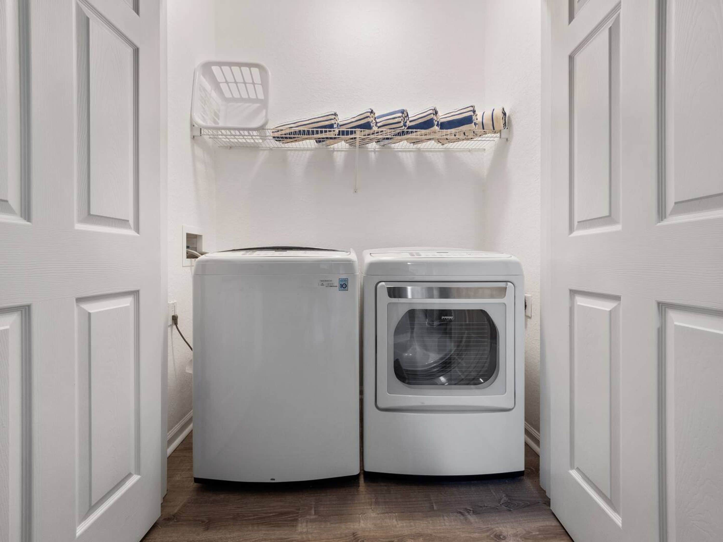 A laundry room with two white appliances, a shelf with folded towels, and a white door.
