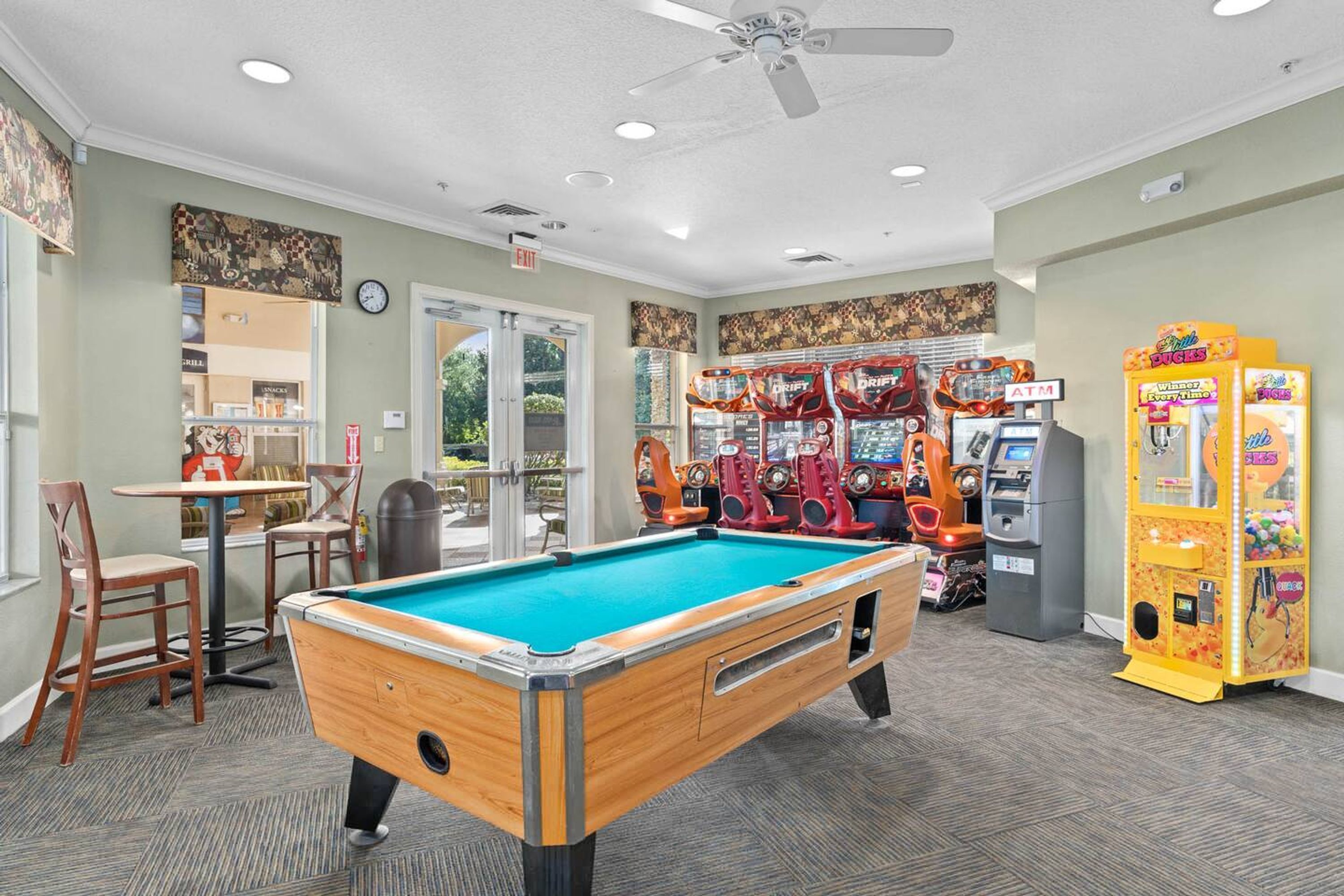 Une table de billard, des jeux d’arcade et un distributeur automatique dans une pièce dotée d’un ventilateur de plafond et d’un sol recouvert de moquette.