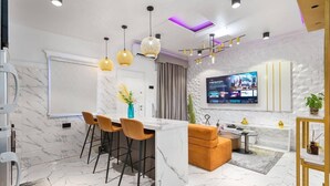 A modern kitchen with a marble countertop, bar stools, pendant lights, and a TV mounted on the wall. - Lekki Grand Apartment (Lekki)