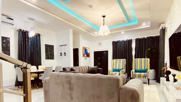 A modern living room with a sofa, chairs, a dining area, and a staircase. - Lekki Grand Apartment (Lekki)