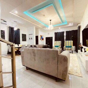 A modern living room with a sofa, chairs, a dining area, and a staircase. - Lekki Grand Apartment (Lekki)