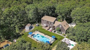 Une maison entourée d’une forêt dense, avec une piscine et un petit abri de jardin.