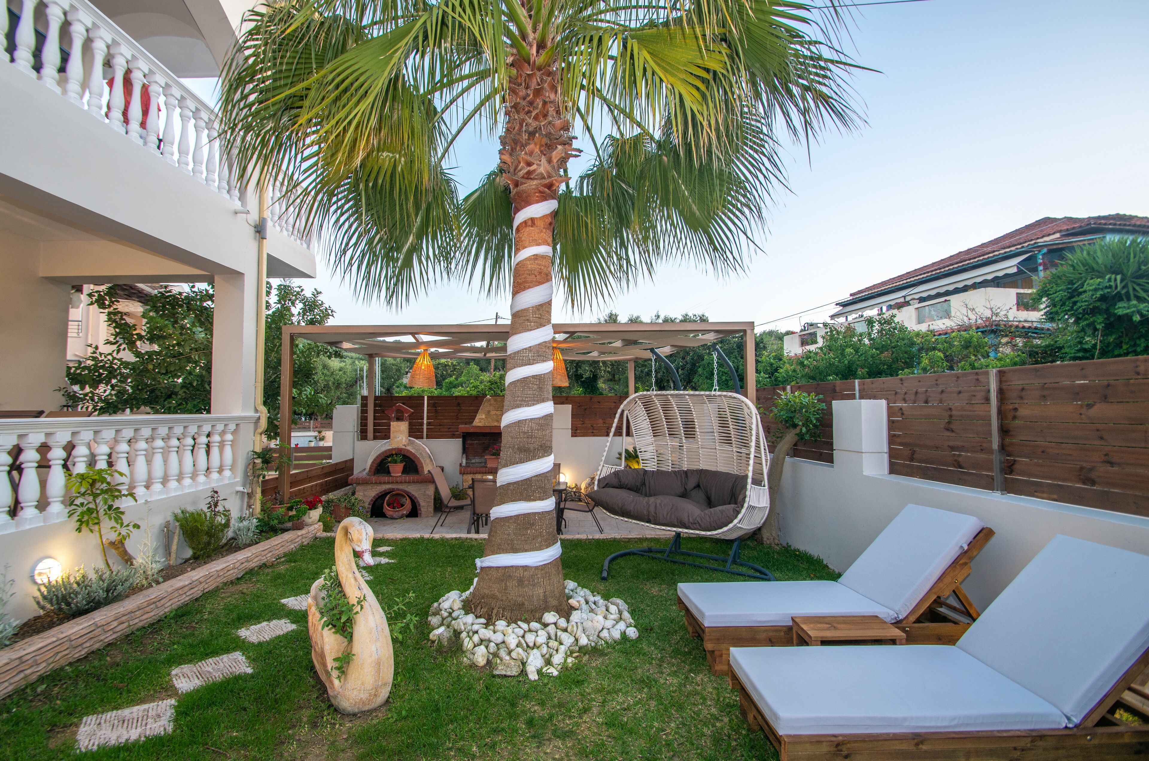Un jardin avec un palmier, une sculpture de cygne, un fauteuil suspendu et deux chaises longues.