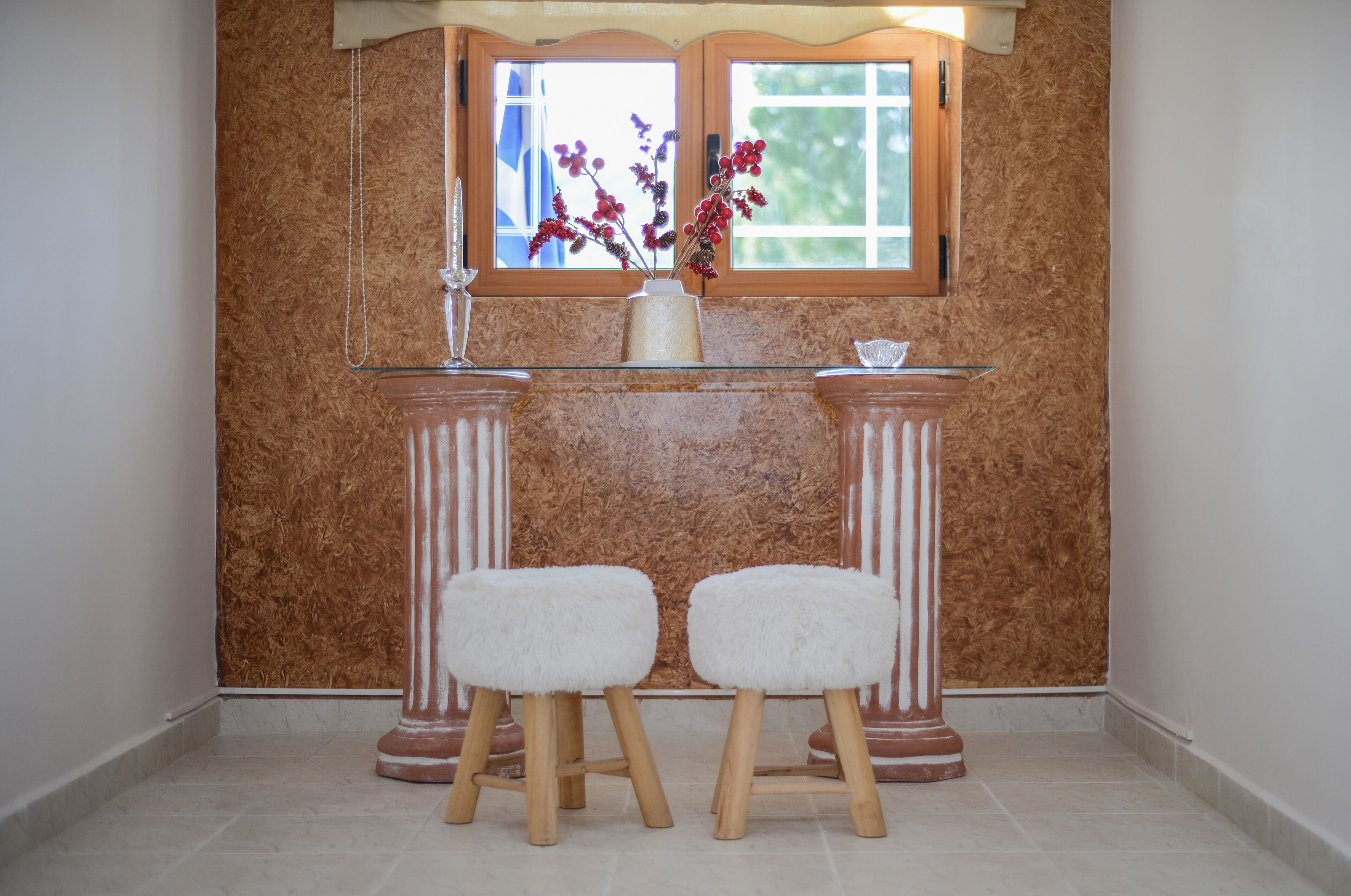 A room with a wooden wall panel, a window with a view, a vase with flowers, and two stools.