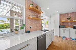 A modern kitchen with white cabinetry, a herringbone-patterned backsplash, wooden shelves, and a view of a swimming pool. - 3 BD Home in Soso Sunny Vista by Rovetravel (West Palm Beach)