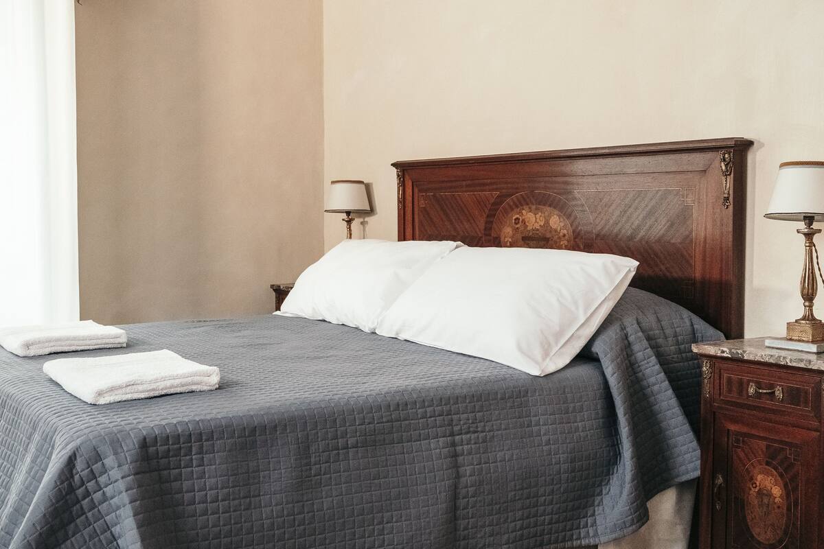 Un lit bien fait, avec une couverture matelassée grise et des oreillers blancs, une table de chevet en bois avec une lampe, et une tête de lit en bois ornée d’un panneau décoratif.