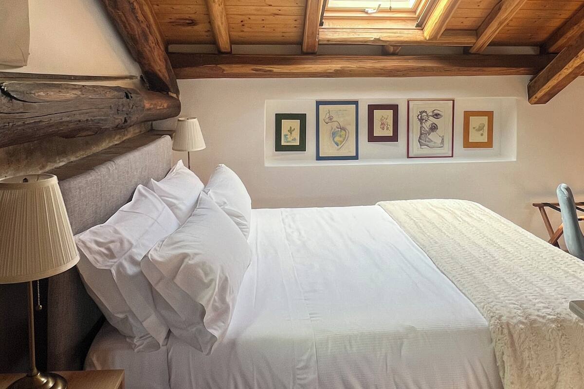 Une chambre avec un plafond en bois, un lit recouvert de draps blancs, une table de chevet avec une lampe et des œuvres d’art encadrées accrochées au mur.