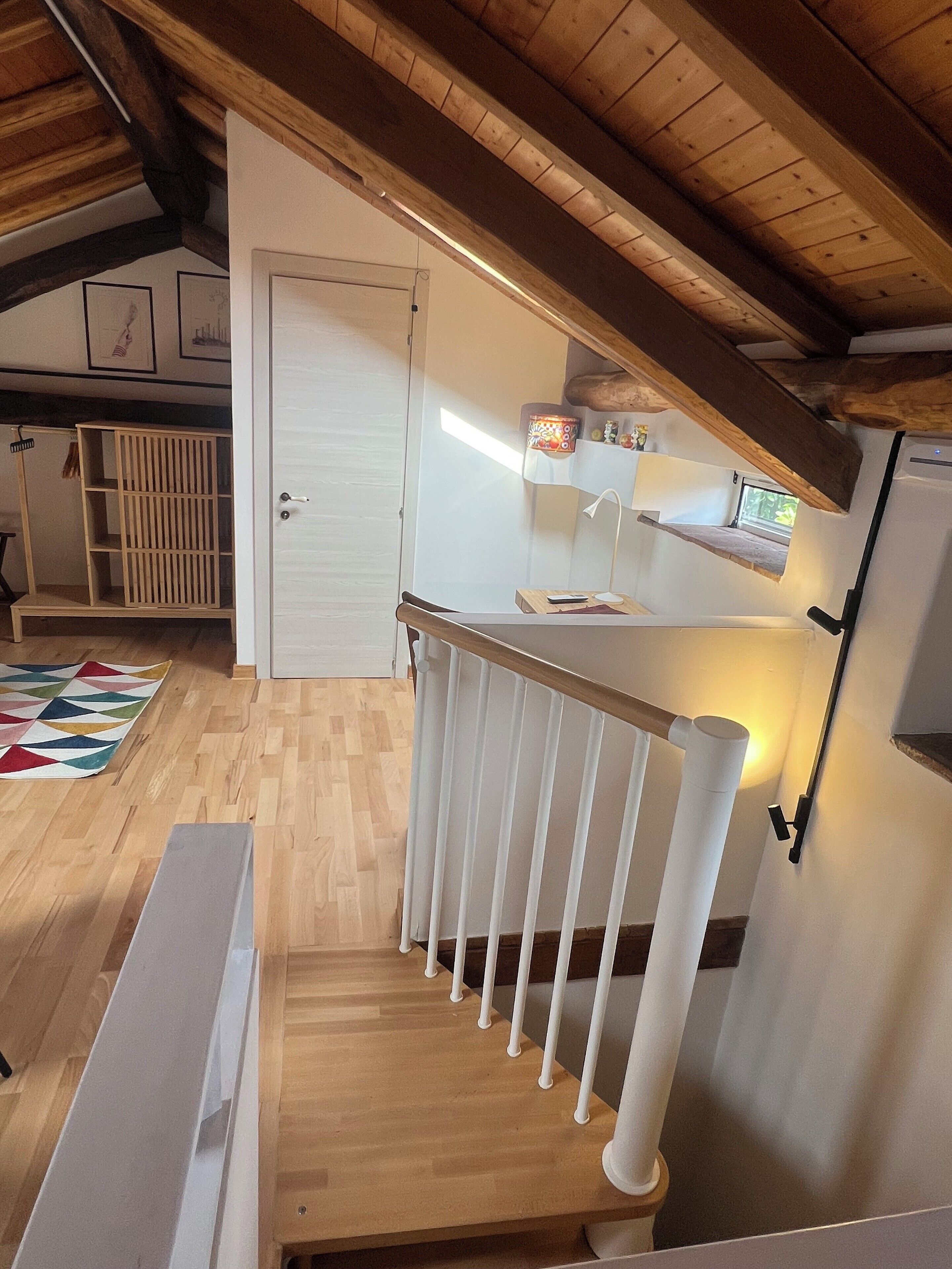 En trappe i træ med hvide gelændere, en hvid dør og et lille køkken med en mikrobølgeovn samt en hylde over.