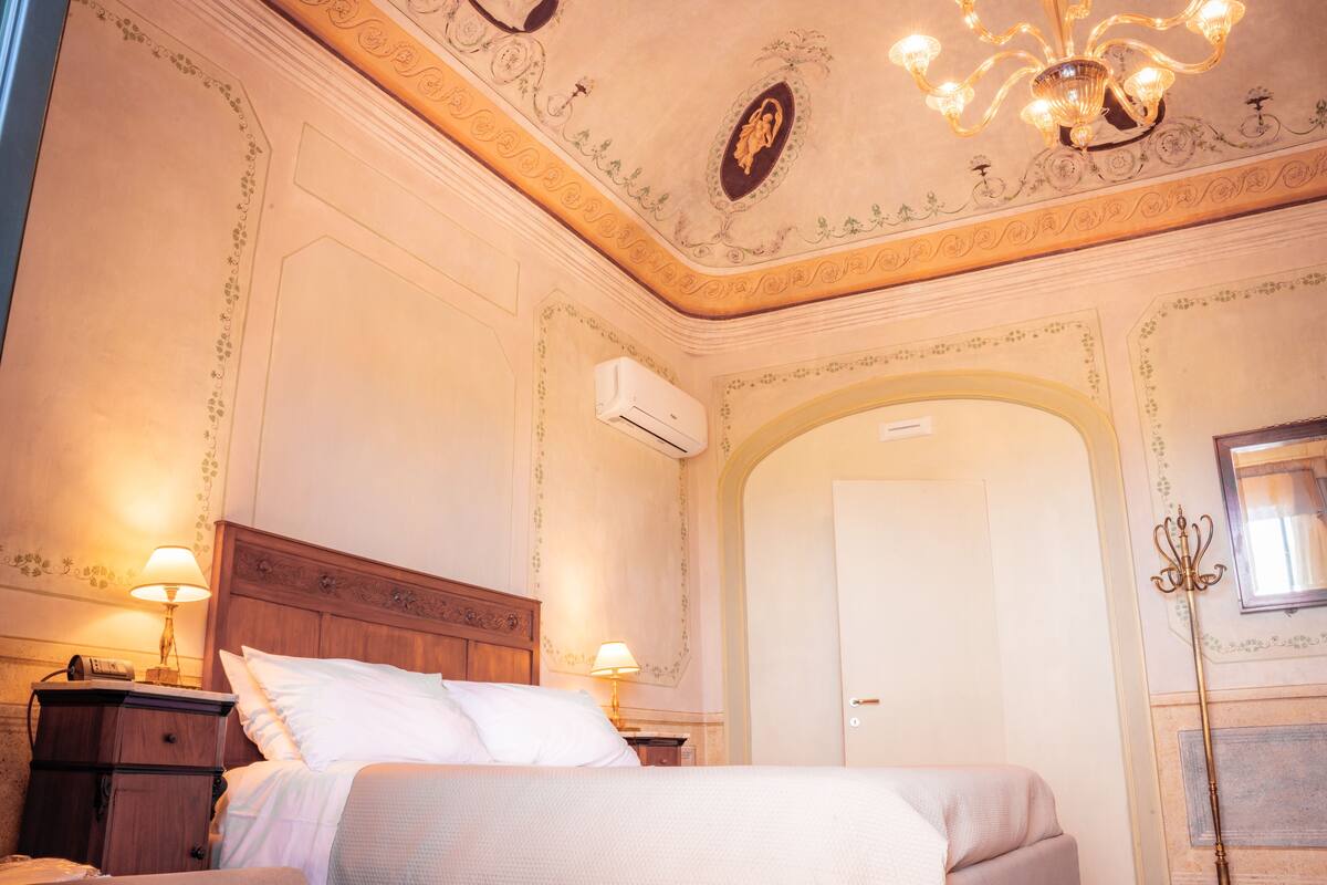 Une chambre d’hôtel avec un lit, deux tables de chevet avec des lampes, une porte en arc de cercle et un lustre.