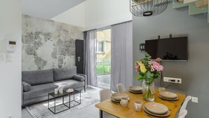Un salon moderne avec un canapé gris, une table à manger en bois mise avec des assiettes et des tasses, un vase rempli de fleurs et un téléviseur fixé au mur.