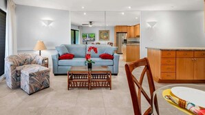 Una sala moderna con sofá, mesa de centro y sillón estampado. Se observa una zona de cocina con armarios de madera y una mesa de comedor.