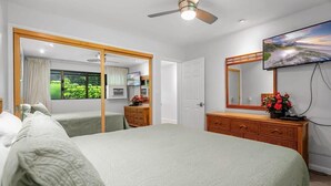 A bedroom with a bed, a dresser, a television, and a window with a view of greenery. - WBH D225 Aloha Ola2 – Relax in Oceanfront Paradise (Kihei)