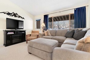 Una sala de estar con televisor de pantalla plana, un sofá, un reposapiés, una silla y una ventana con cortinas.