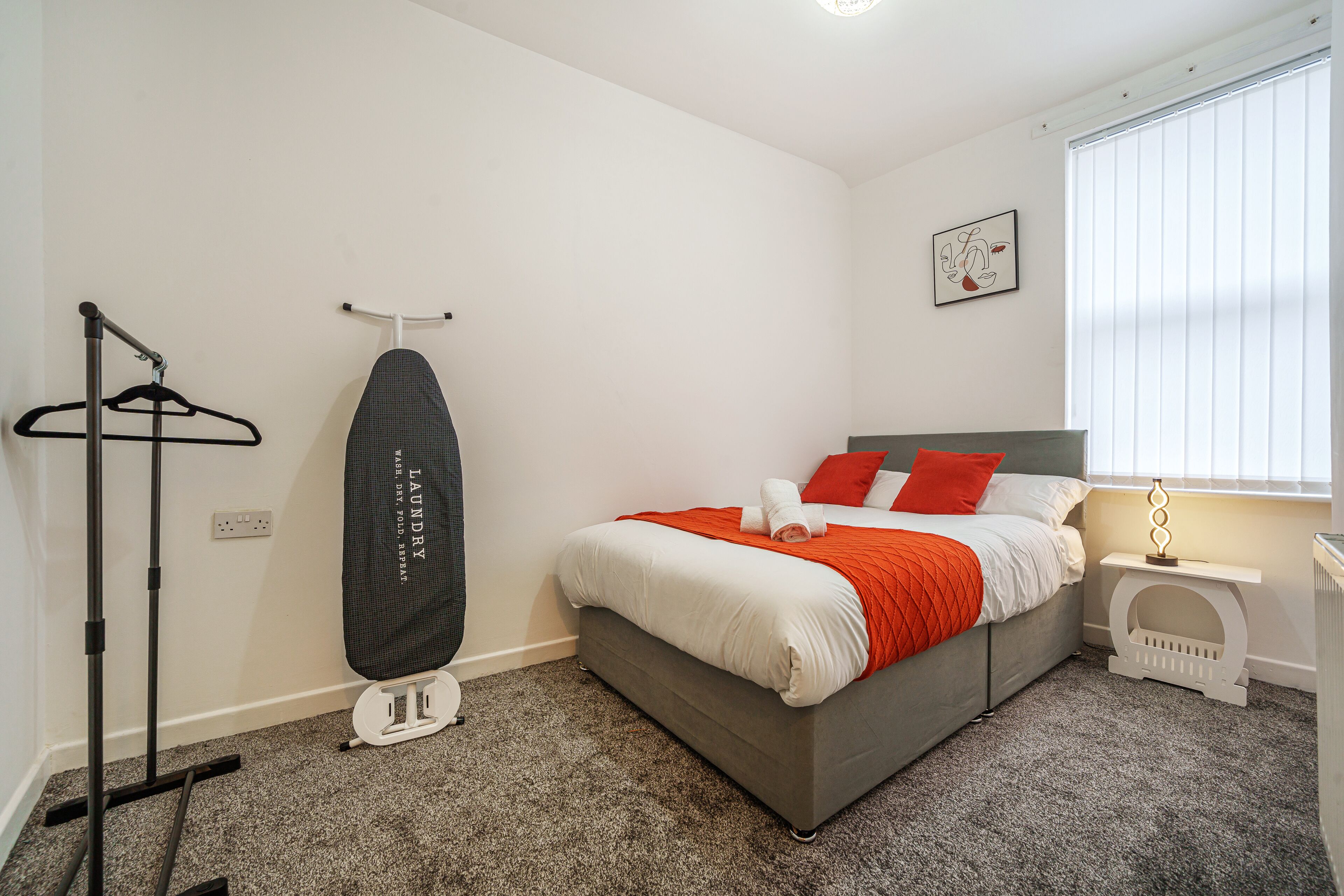 A neatly arranged bedroom with a bed, a hanging laundry rack, a radiator, and a window with blinds.