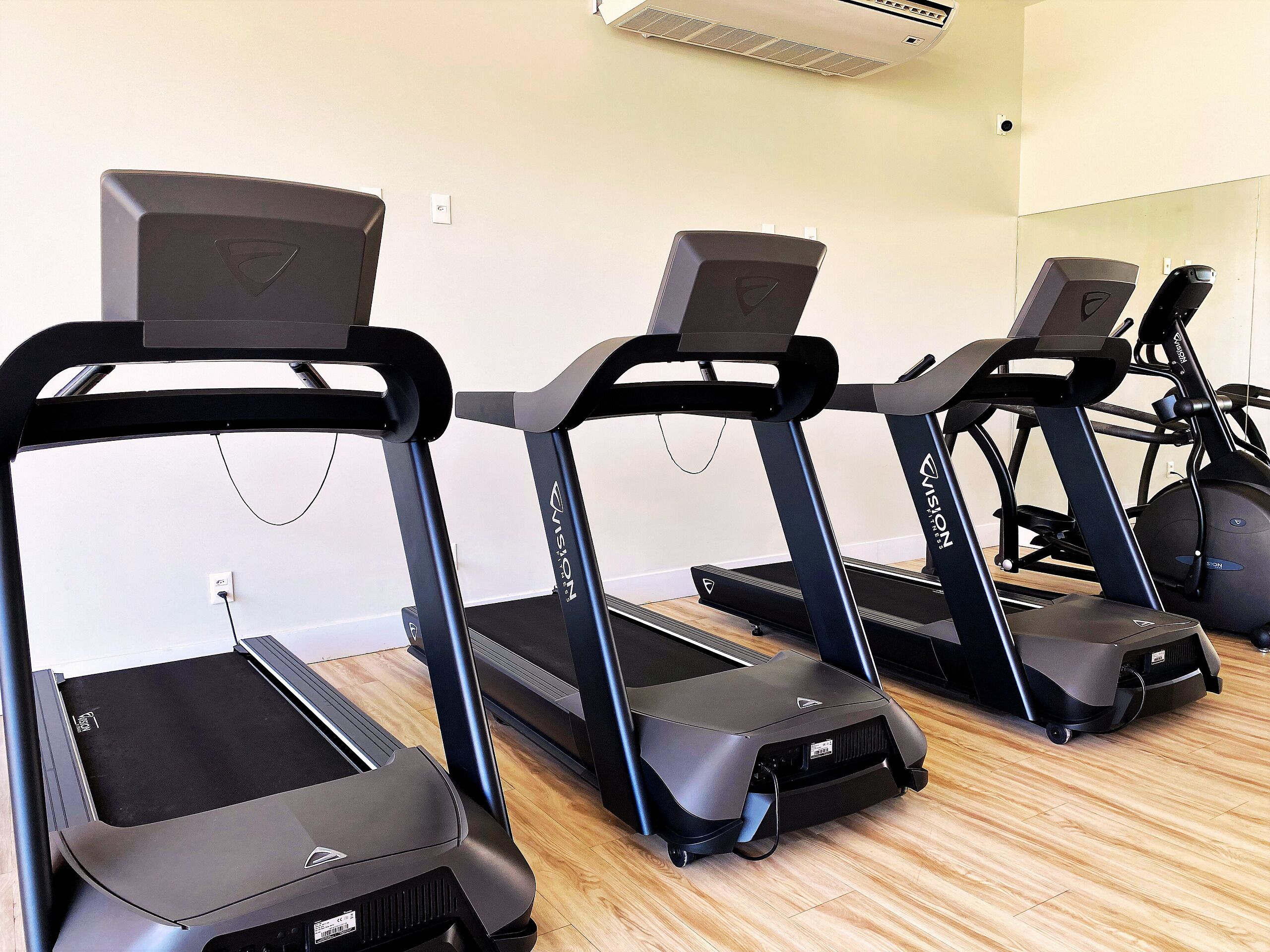 Varias cintas de correr estacionadas en un gimnasio con suelo de madera y pared blanca.