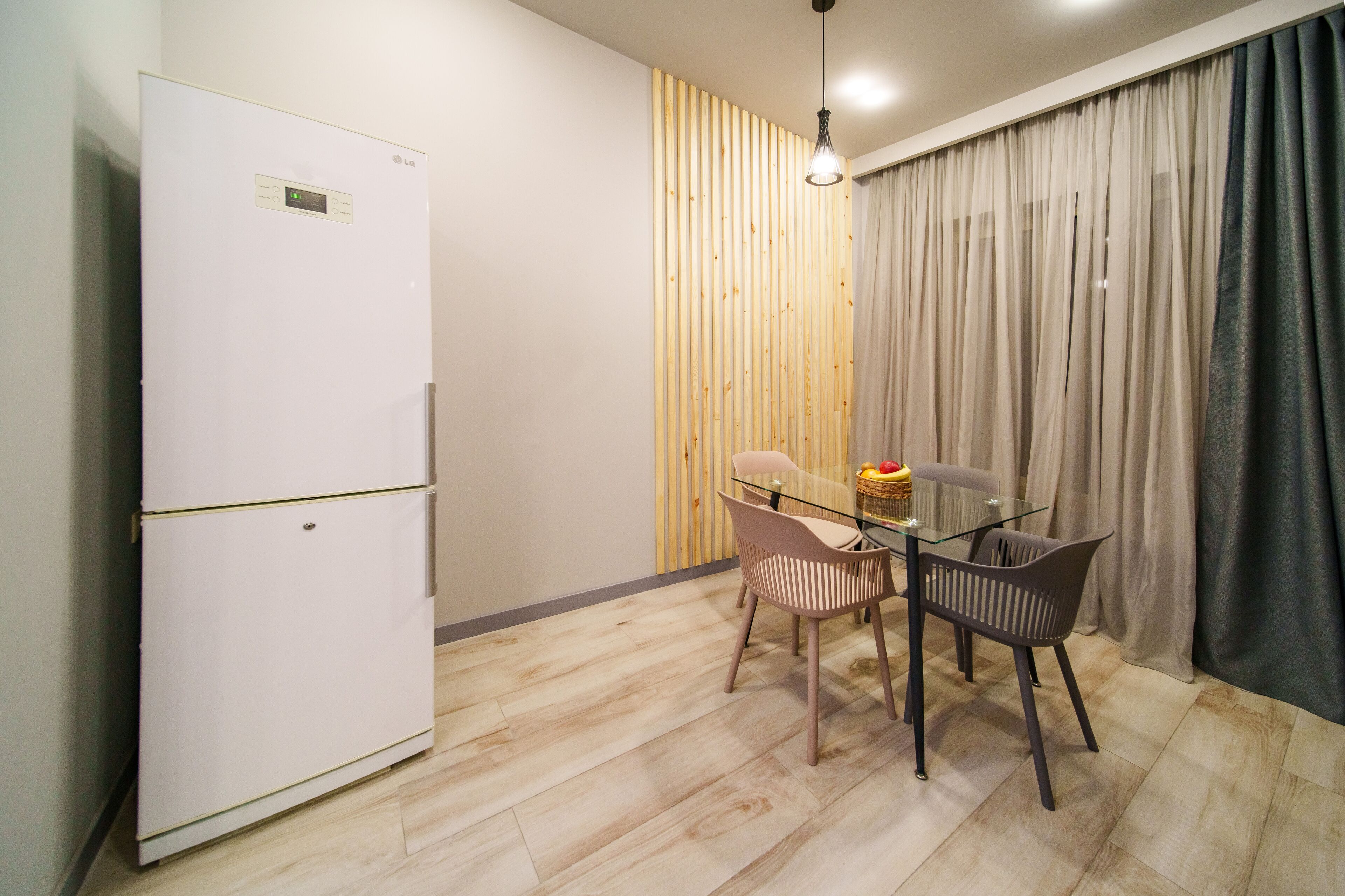 A compact kitchen with a white refrigerator, a wooden accent wall, a glass dining table with two chairs, and a large window with sheer curtains.