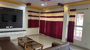 A living room with a flat-screen TV mounted on the wall, a brown leather sofa, a glass-top coffee table, and red and beige curtains. - 2 BHK Banglow (Mahabaleswer)