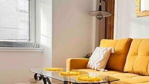 A living room with a yellow sofa, a glass coffee table with yellow objects, and a window with blinds. - 1-bed Apartment Sleeps 2 -free Parking -wifi & Tv (Dundee)
