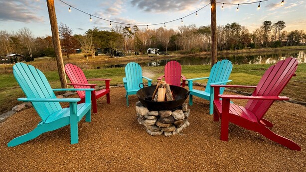 A fire pit with logs surrounded by four Adirondack chairs, set on gravel with a lake and trees in the background. - Lake Therapy at The Retreat at Water's Edge (Tracy City)
