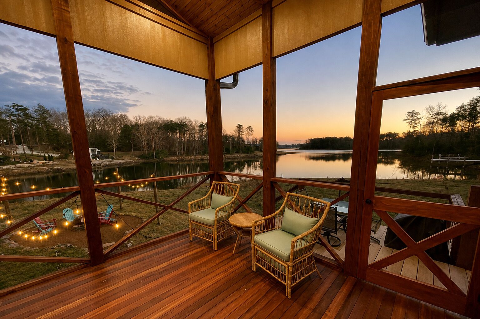 A wooden deck with wicker chairs and a table, overlooking a calm lake and trees.