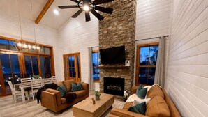 A spacious living room with a stone fireplace, wooden ceiling beams, and a large window overlooking a snowy landscape. - Lake Therapy at The Retreat at Water's Edge (Tracy City)