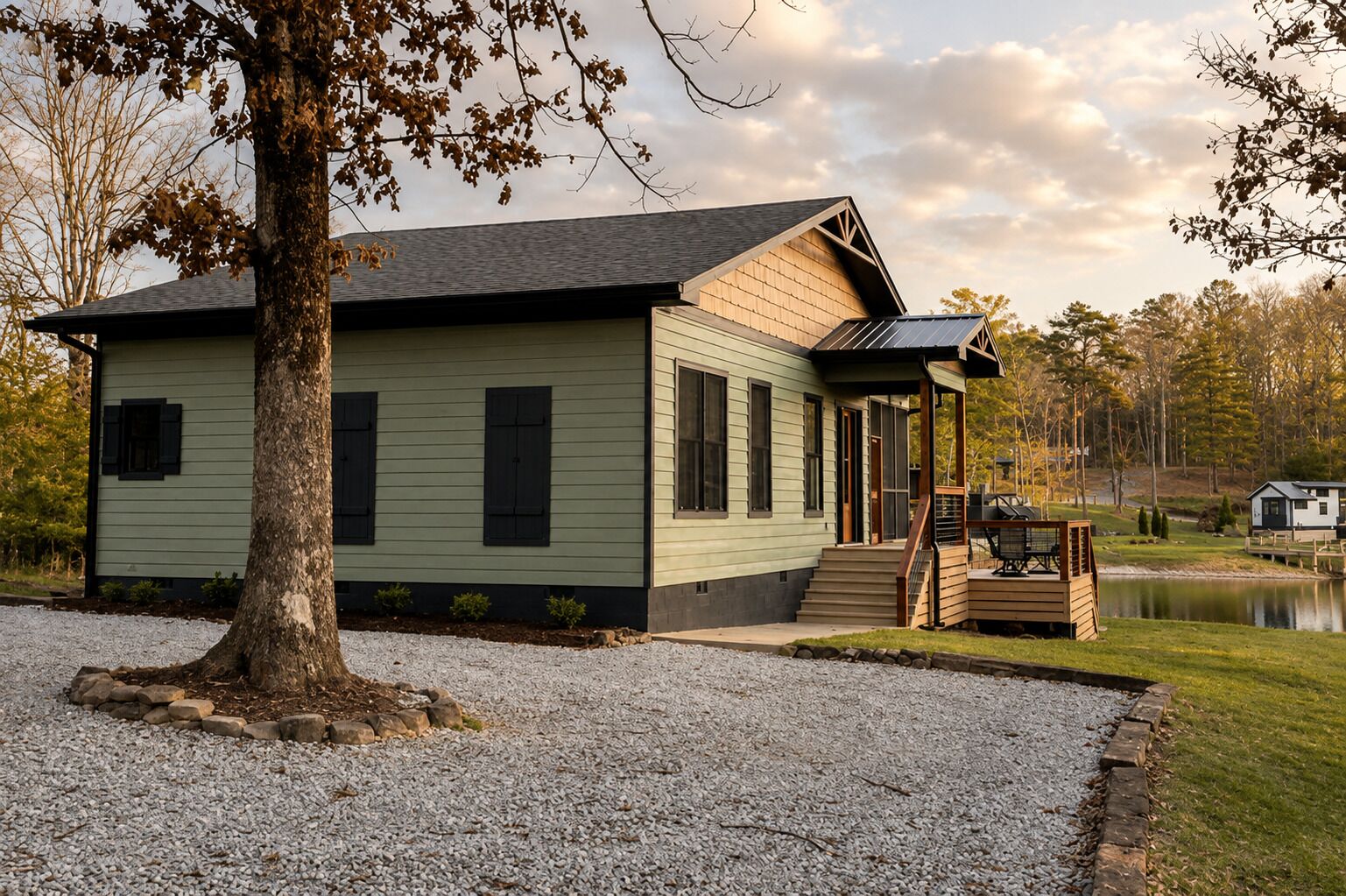 Une cabane de plain-pied dotée d’un porche couvert et d’une petite terrasse, entourée d’une allée en gravier et d’un étang.