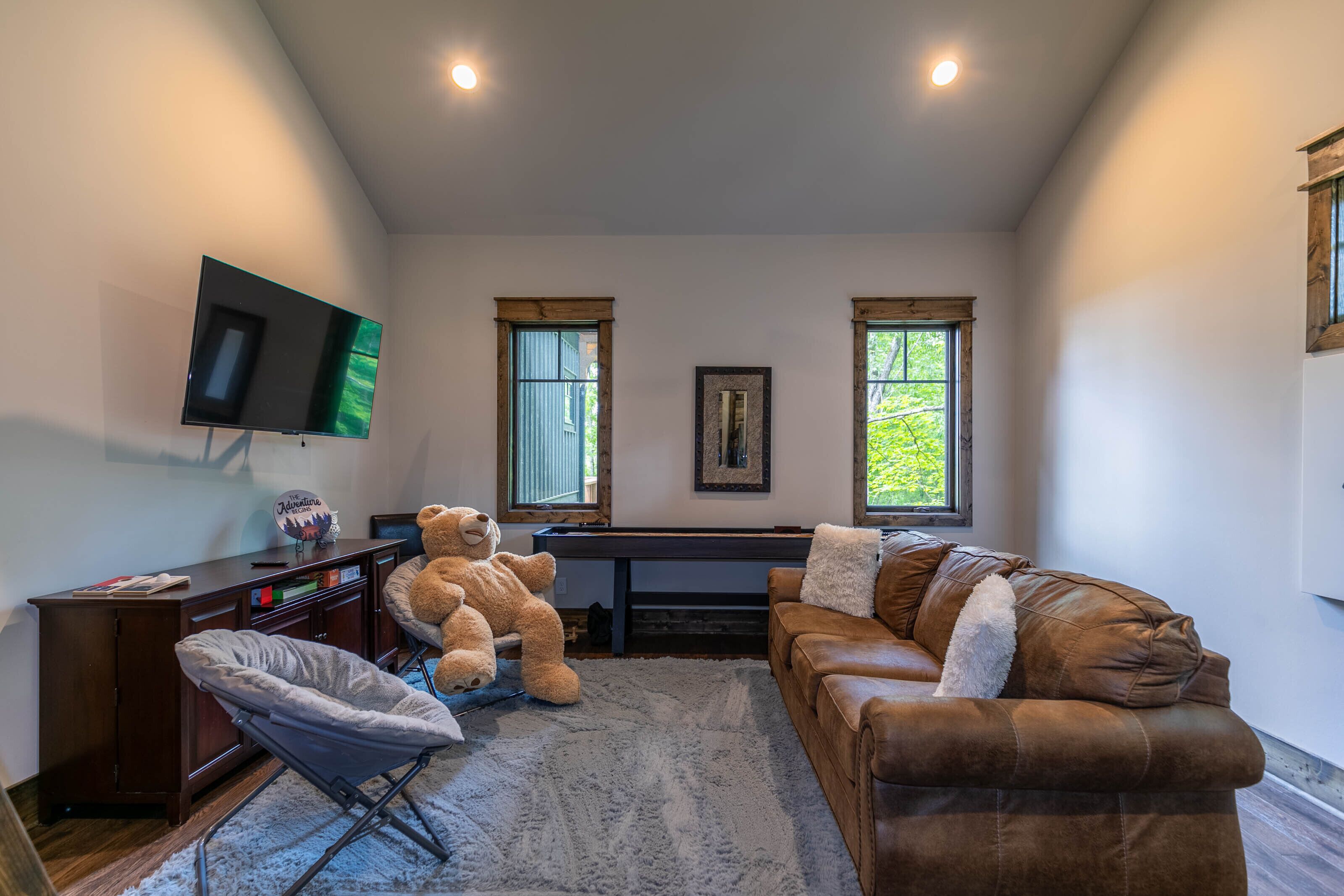 Una sala de estar con un televisor de pantalla plana grande, un sofá de cuero marrón, una silla pequeña y un escritorio con un osito de peluche.