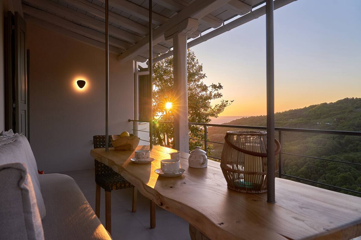 A balcony with a wooden table, a wicker chair, and a view of the sunset over a hilly landscape.