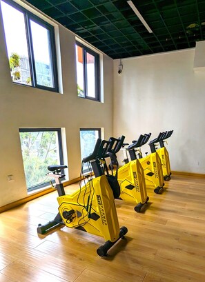 A room with four stationary bikes arranged in a row, large windows with views of greenery, and a wooden floor.
