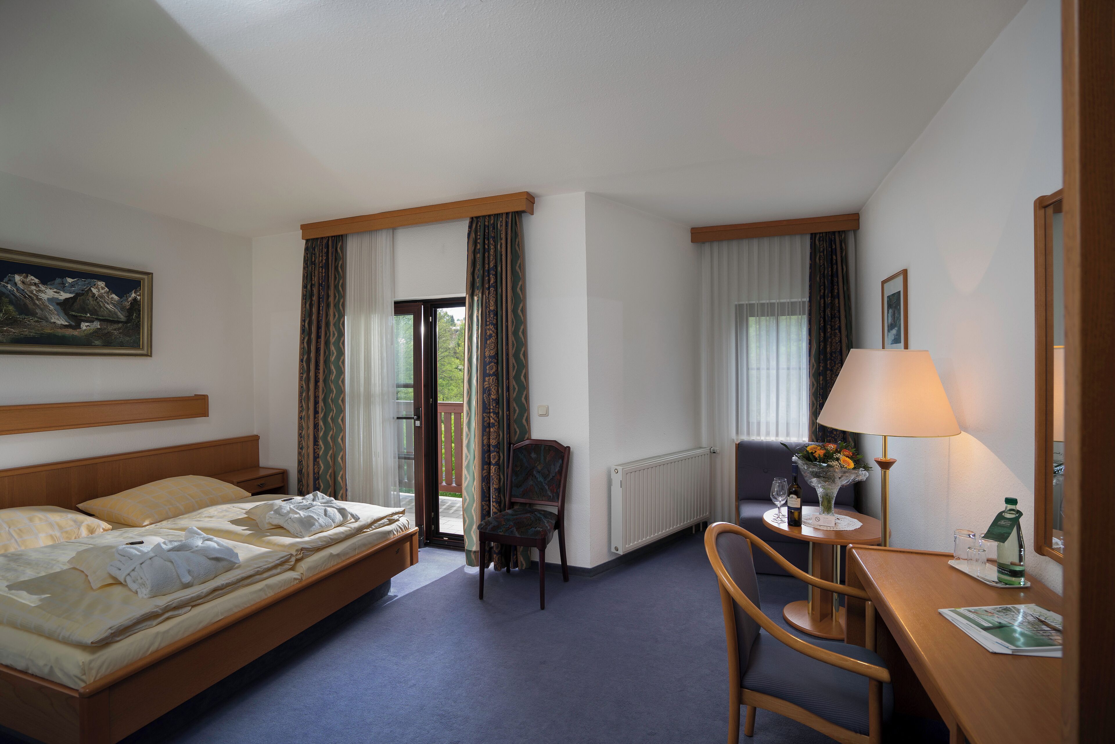 A hotel room with a bed, a desk, a chair, a window with curtains, a radiator, and a painting on the wall.