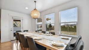 A dining area with a long table set for four, a chandelier, and large windows with blinds.