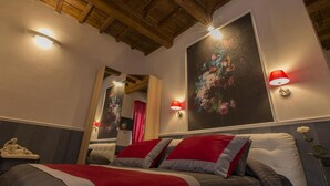 A hotel room with a wooden ceiling, a large bed with red and grey bedding, two wall-mounted lamps, and a floral painting. - Suite Apartment 2/4 Persons (Firenze)