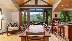 Uma sala de jantar com mesa e cadeiras de madeira, um banco com almofadas e vista para o exterior por grandes janelas.