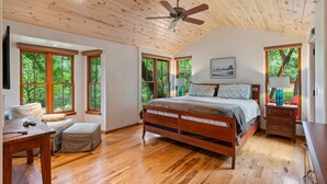 Um quarto espaçoso com teto de madeira, janelas grandes, uma cama, uma escrivaninha e uma cadeira.