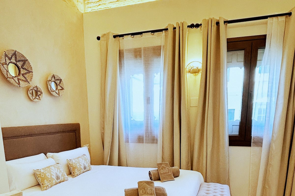 Una habitación de hotel con una cama grande, un banco, una mesita de noche y una ventana con cortinas.