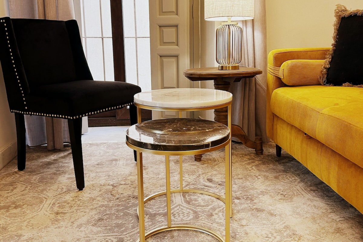 Una habitación con un sillón tapizado en negro, una mesa redonda con tapa de mármol, un sofá amarillo y una lámpara sobre una mesita.