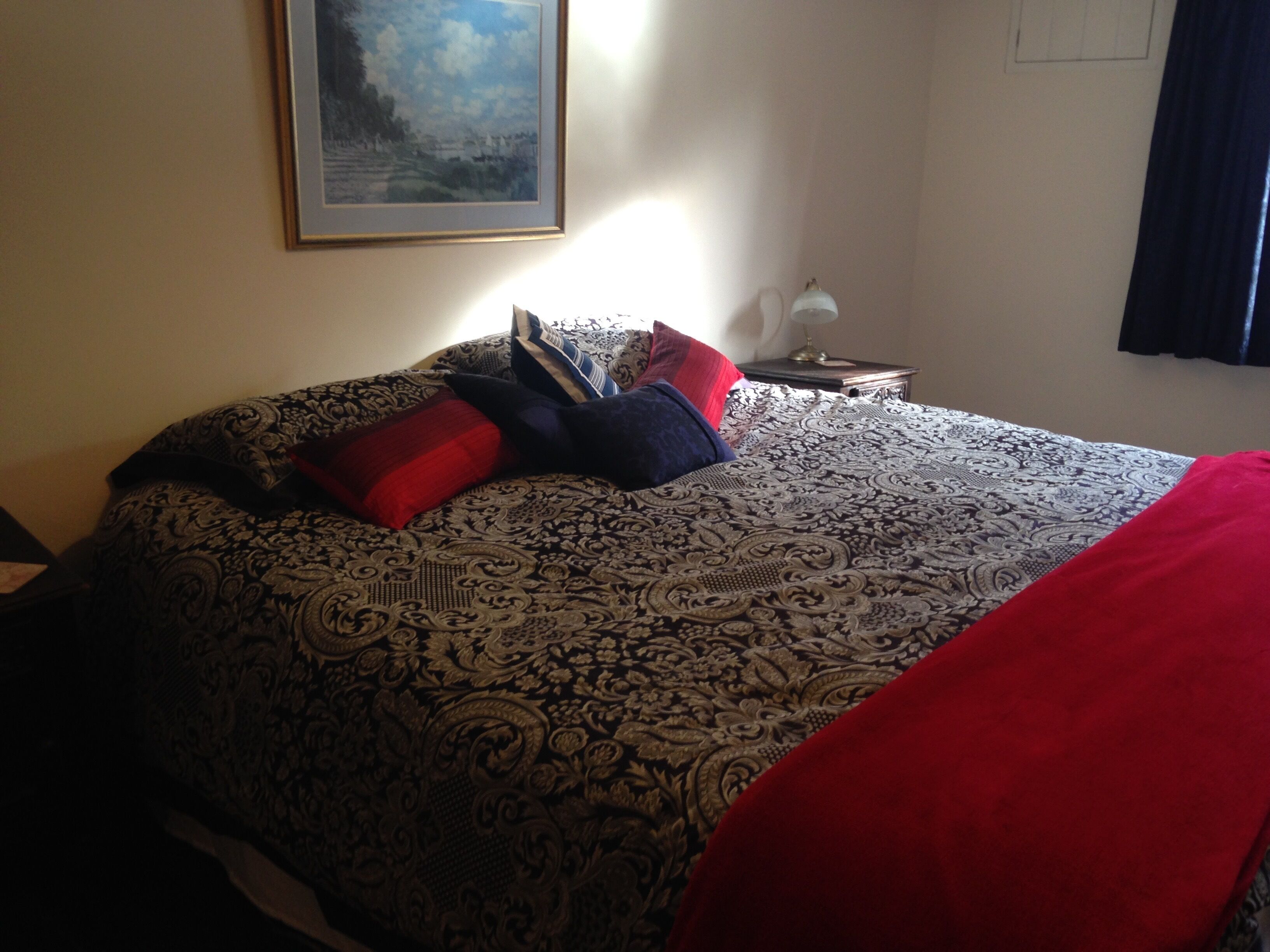 A bedroom with a patterned bedspread, a red throw blanket, and a framed painting on the wall.
