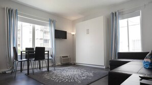 En moderne stue med sofa, spisebord med stole, et fladskærms TV monteret på væggen og et stort vindue med gardiner.