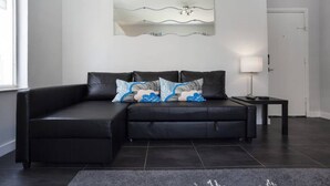 En moderne stue med en sort lædersofa, en dekorativ vægdekoration, et lille bord med en lampe og en mørk flisegulv.