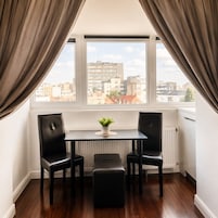 A corner room with a table, two chairs, a bench, a radiator, and a window with curtains.