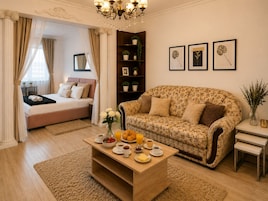 A bedroom with a bed, a sitting area with a sofa, a coffee table with breakfast items, and a bookshelf with plants.