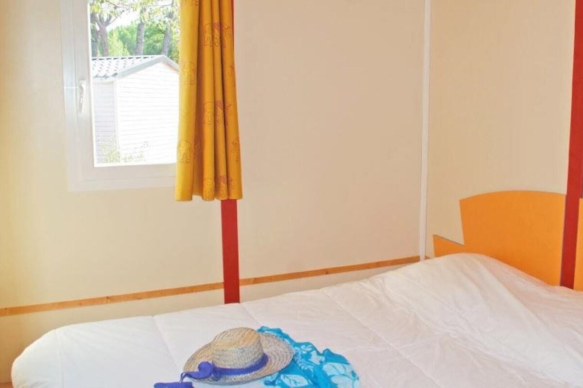 A single bed with an orange headboard in a room with plain walls and a window with yellow curtains.