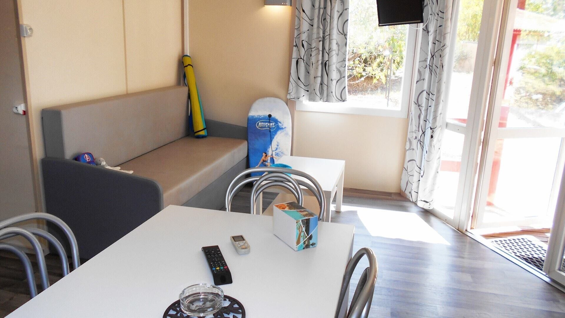 A room with a white table, a bench, a surfboard, and a window with curtains.