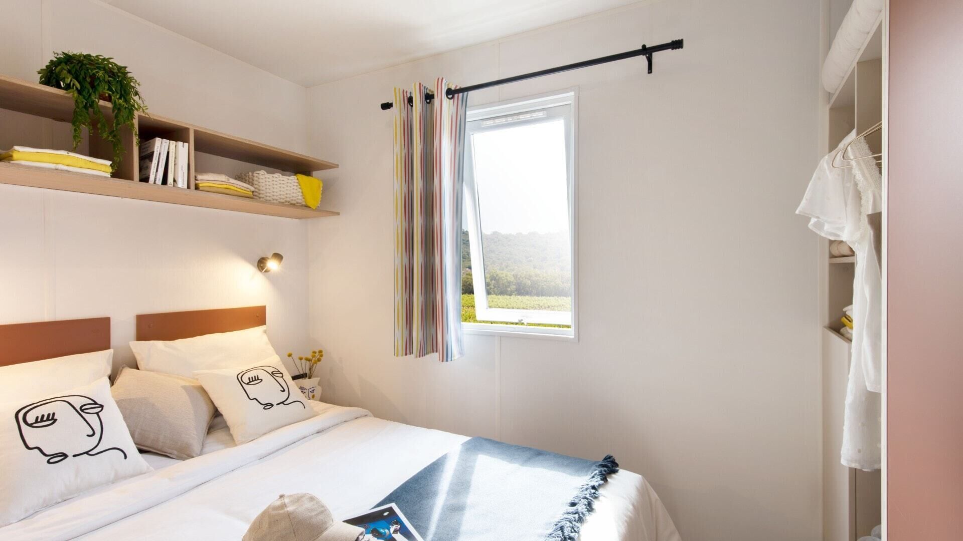 A bedroom with a bed, pillows, a bedside table, a window with curtains, a shelf with books and a plant, and a wardrobe with clothes.