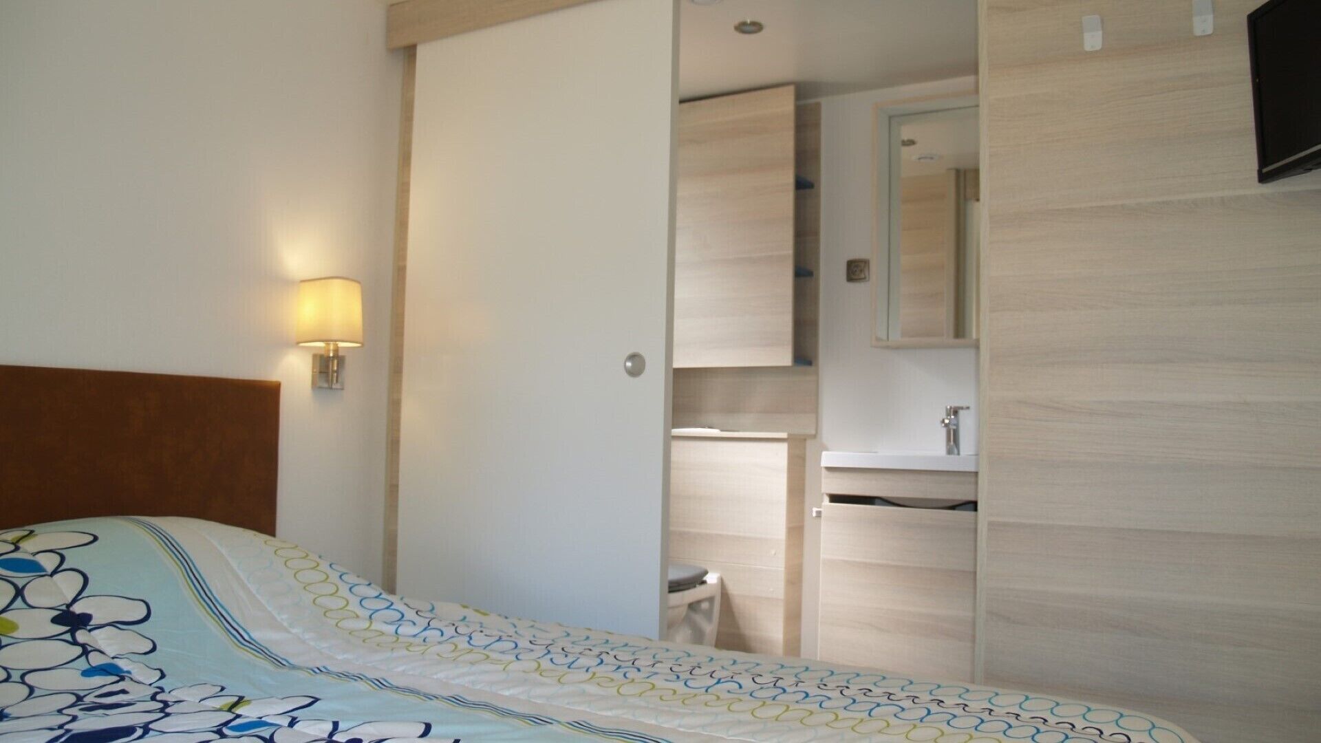 A hotel room with a bed, a nightstand, a wall-mounted lamp, a bathroom with a sink and mirror, and a wooden door.