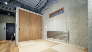 Et rum med stråmåtter, en skydedør i træ, en bænk og en ventilationsrist på væggen.
