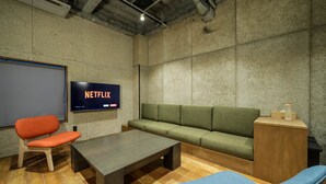 Et mødelokale med et stort skærm, der viser 'NETFLIX', en grøn sofa, et træbord og en blå stol.