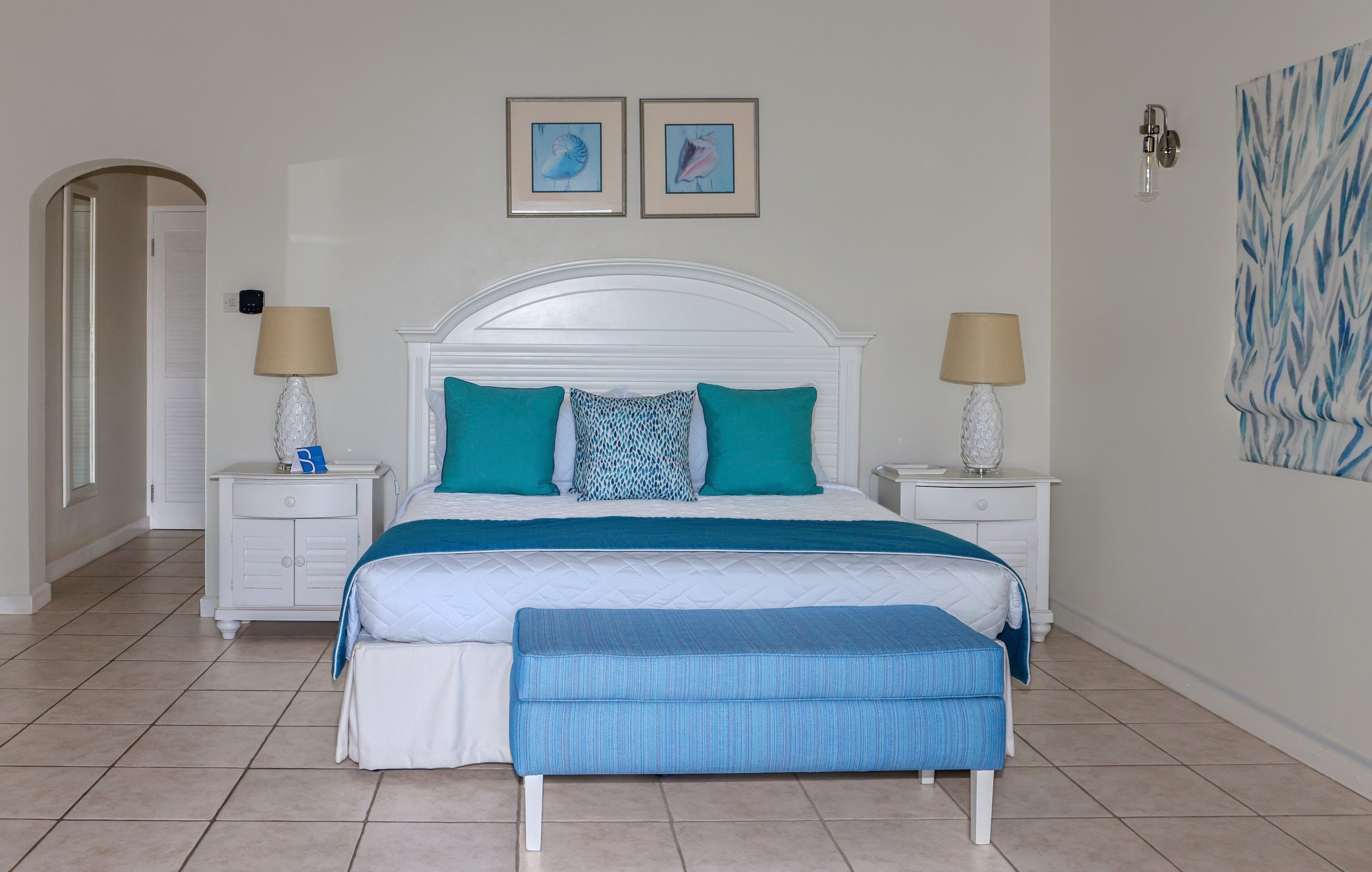 Une chambre avec un lit blanc, des oreillers bleu turquoise et blancs, un banc bleu, deux tables de chevet avec des lampes et une porte donnant sur une autre pièce.