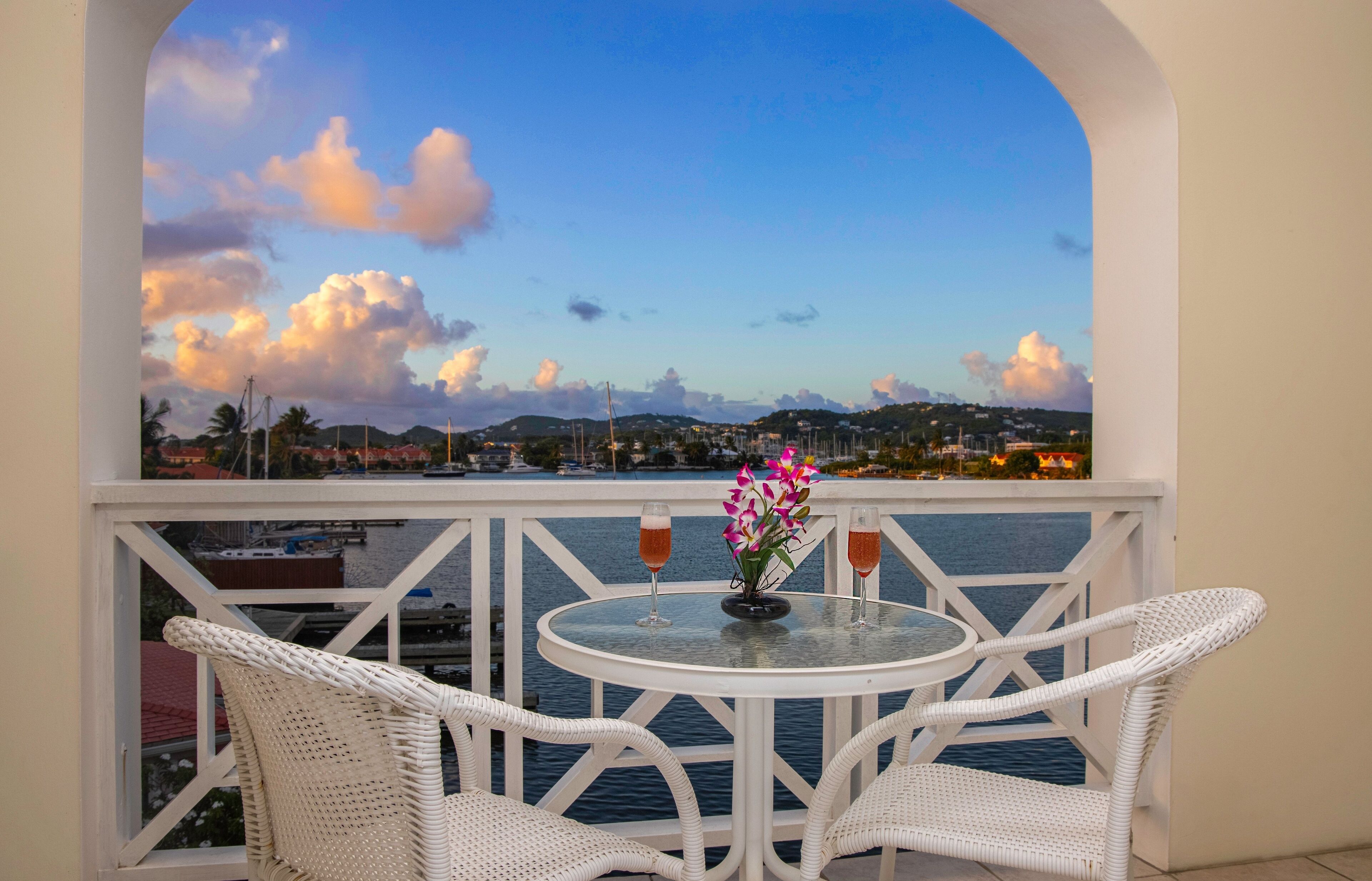 A balcony with white wicker chairs, a glass table, and a vase with flowers.